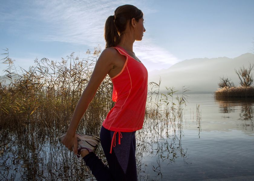A woman connects with nature, stretching by a lake at sunrise to build fitness and endurance with support from E3Live.