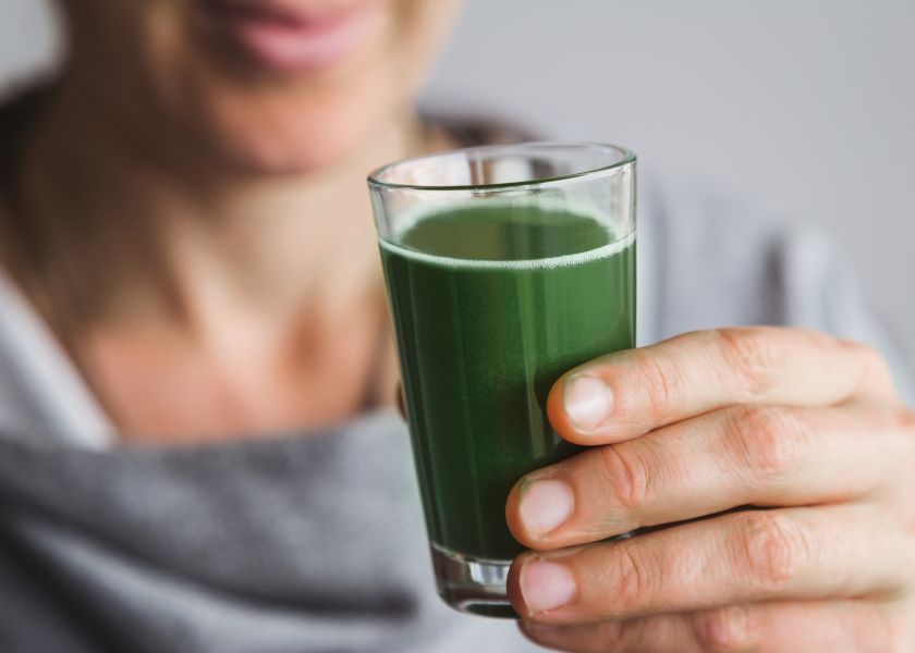 A person holds a small glass filled with a vibrant green E3AFA® or E3Live® drink. The rich, deep green liquid suggests a concentrated shot of fresh-frozen Aphanizomenon flos-aquae (AFA) microalgae. The person is partially out of focus, drawing attention to the nutrient-dense superfood beverage in the foreground, captured in a clean, natural setting.
