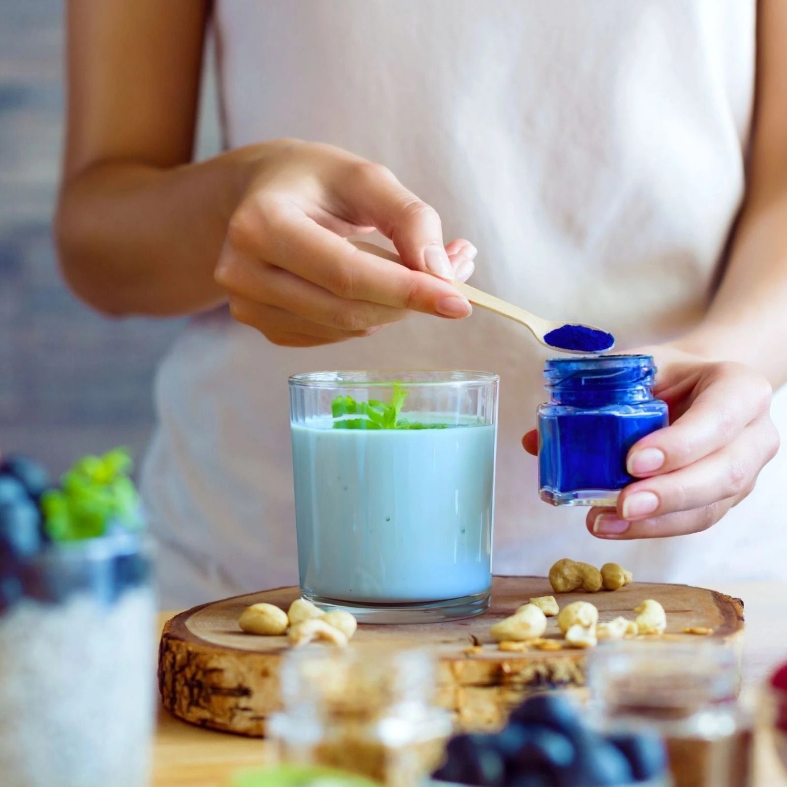 Person adding blue powder to a glass of milk with nuts and berries on a wooden board.