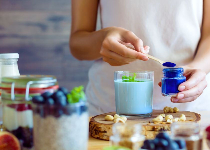 Close-up of a person preparing a vibrant blue smoothie using BlueMAJIK powder. The scene features jars of fresh ingredients like blueberries, cashews, and yogurt in the foreground, with a glass of blue-tinted milk garnished with mint leaves on a rustic wooden tray.