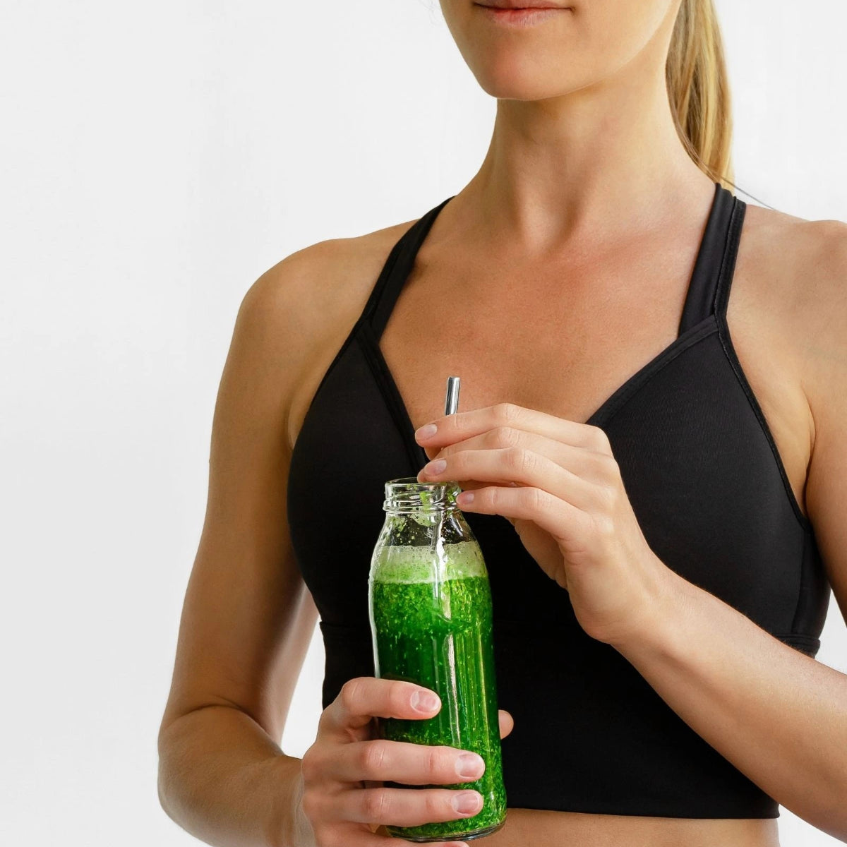Woman holding a green smoothie in a glass bottle with a straw against a white background
