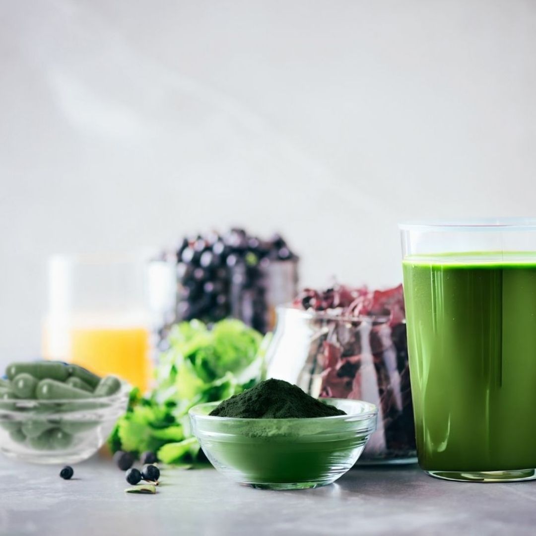 Green smoothie with ingredients including a bowl of green E3AFA powder and a bowl of E3AFA capsules on a light background.