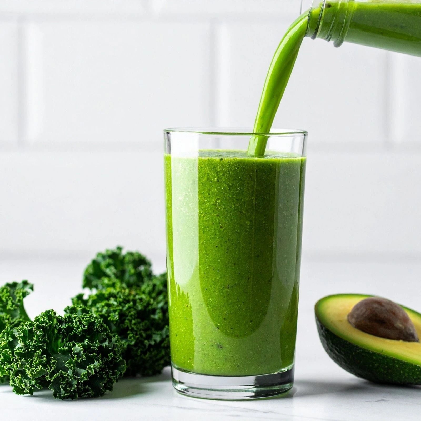 Green smoothie being poured into a glass with kale and avocado on a white background