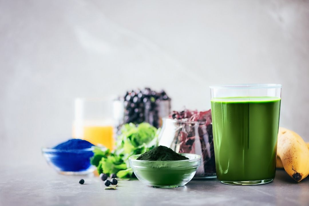 Green smoothie in a glass with ingredients like bananas, berries, and a blue powder on a light background