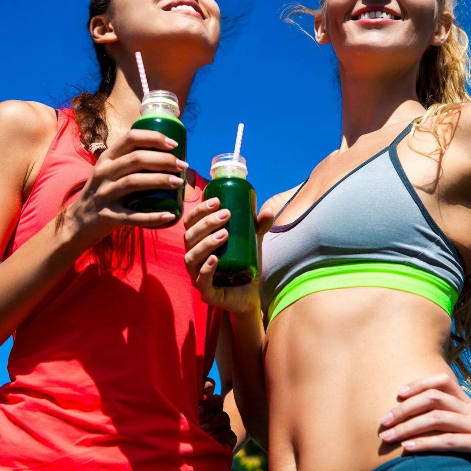 Two women in athletic wear holding green smoothies against a blue sky.