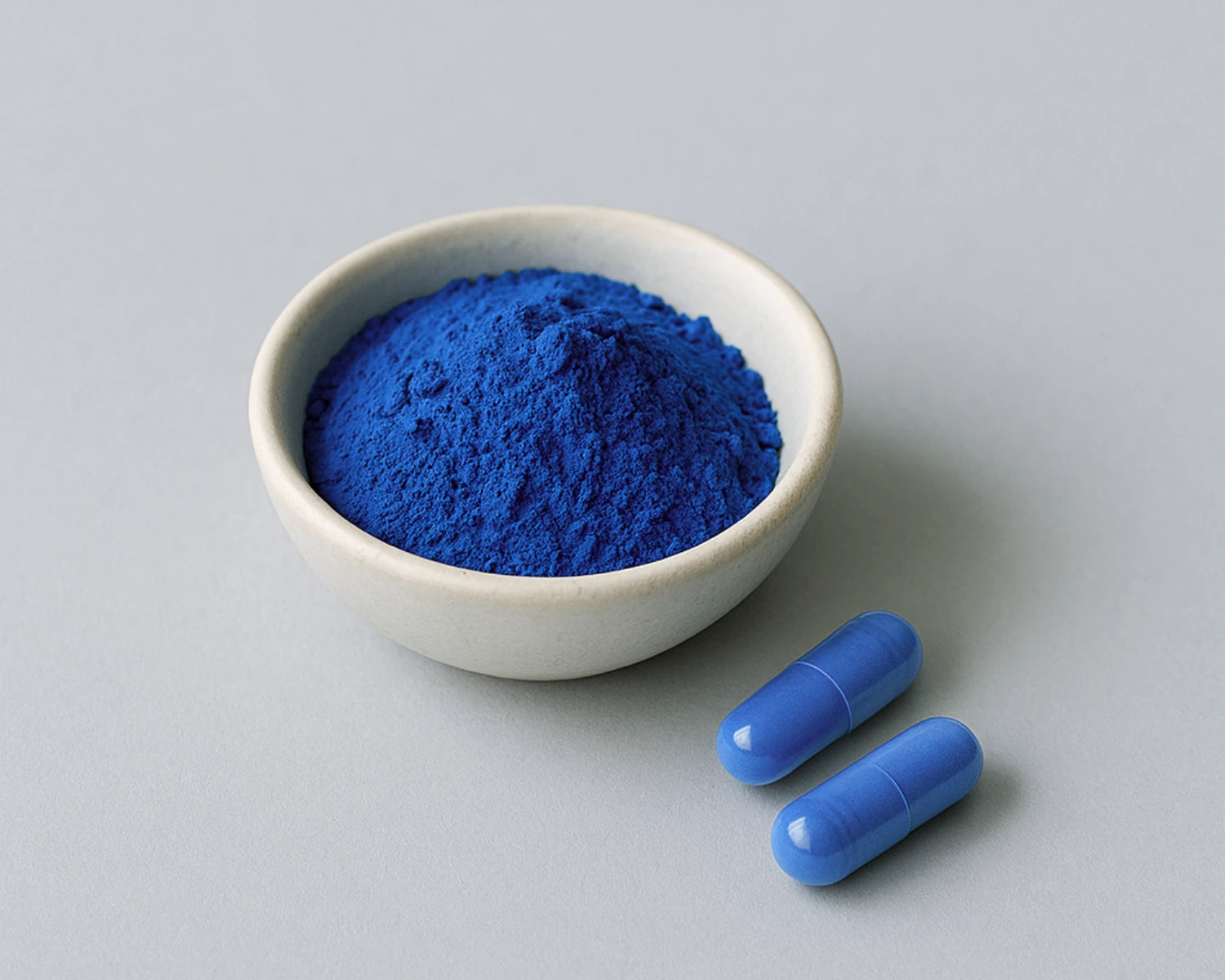 White bowl with blue powder and two blue capsules on a gray background