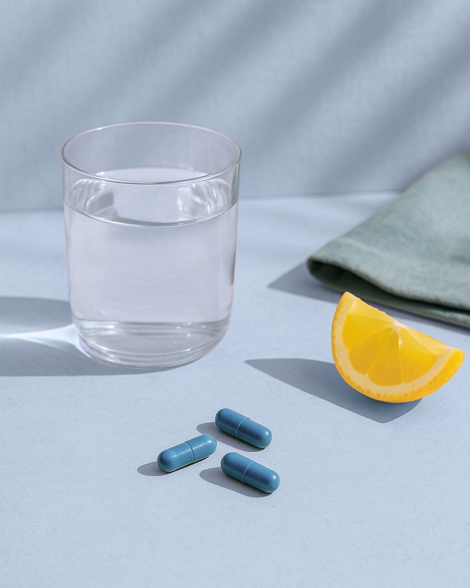 Blue capsules, a glass of water, and a lemon wedge on a light blue surface.