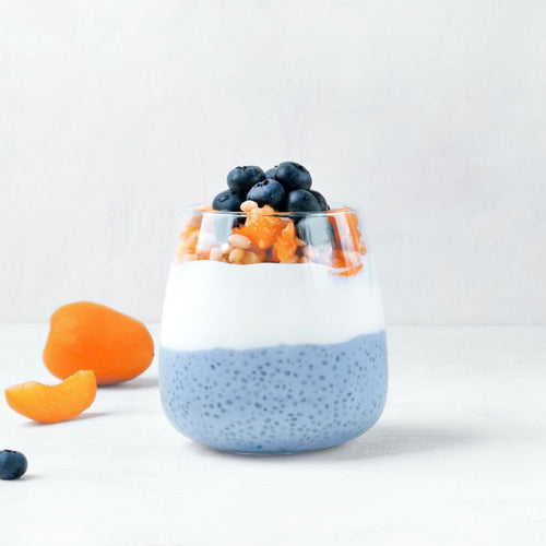 A beautifully layered chia pudding in a clear glass, featuring a blue-tinted chia seed mixture at the bottom, a smooth white yogurt layer in the middle, and a topping of fresh blueberries, diced apricots, and pine nuts. The background is a clean, white surface with whole and sliced apricots scattered around, adding a natural touch. The blue hue suggests the use of BlueMAJIK, a spirulina extract known for its vibrant color and nutritional benefits.