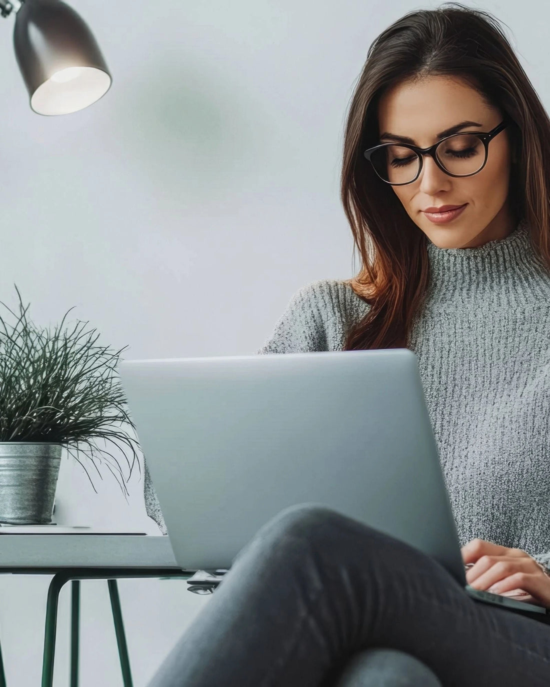 Woman using a laptop in a home office setting with a plant and lamp.