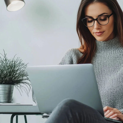 Woman using a laptop in a home office setting with a plant and lamp.
