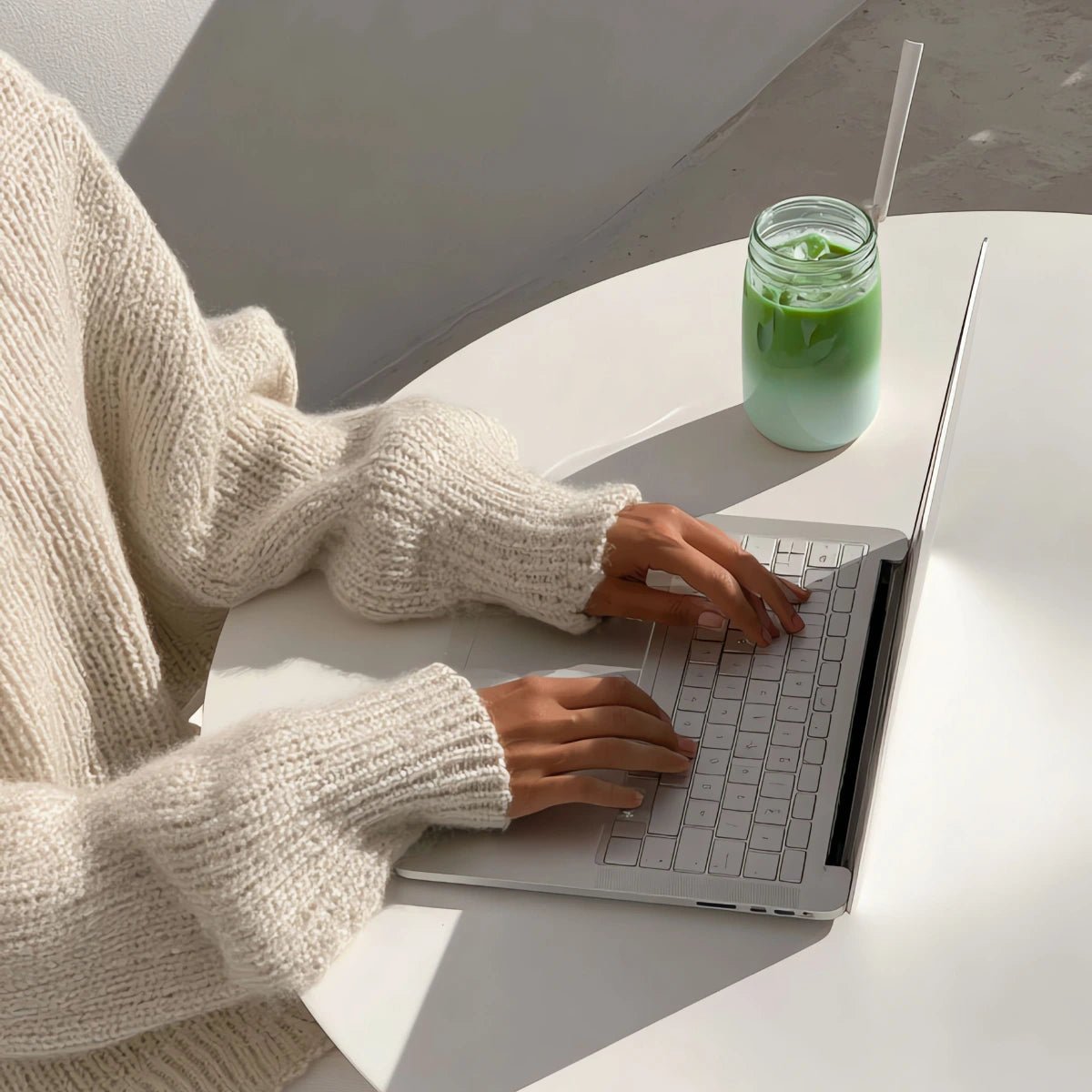 Person using a laptop with a green drink on a white surface