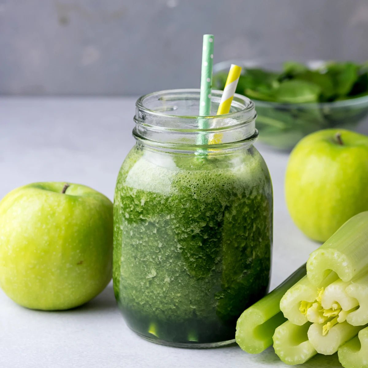 Green smoothie in a jar with a straw, surrounded by green apples, celery, and spinach on a light surface.