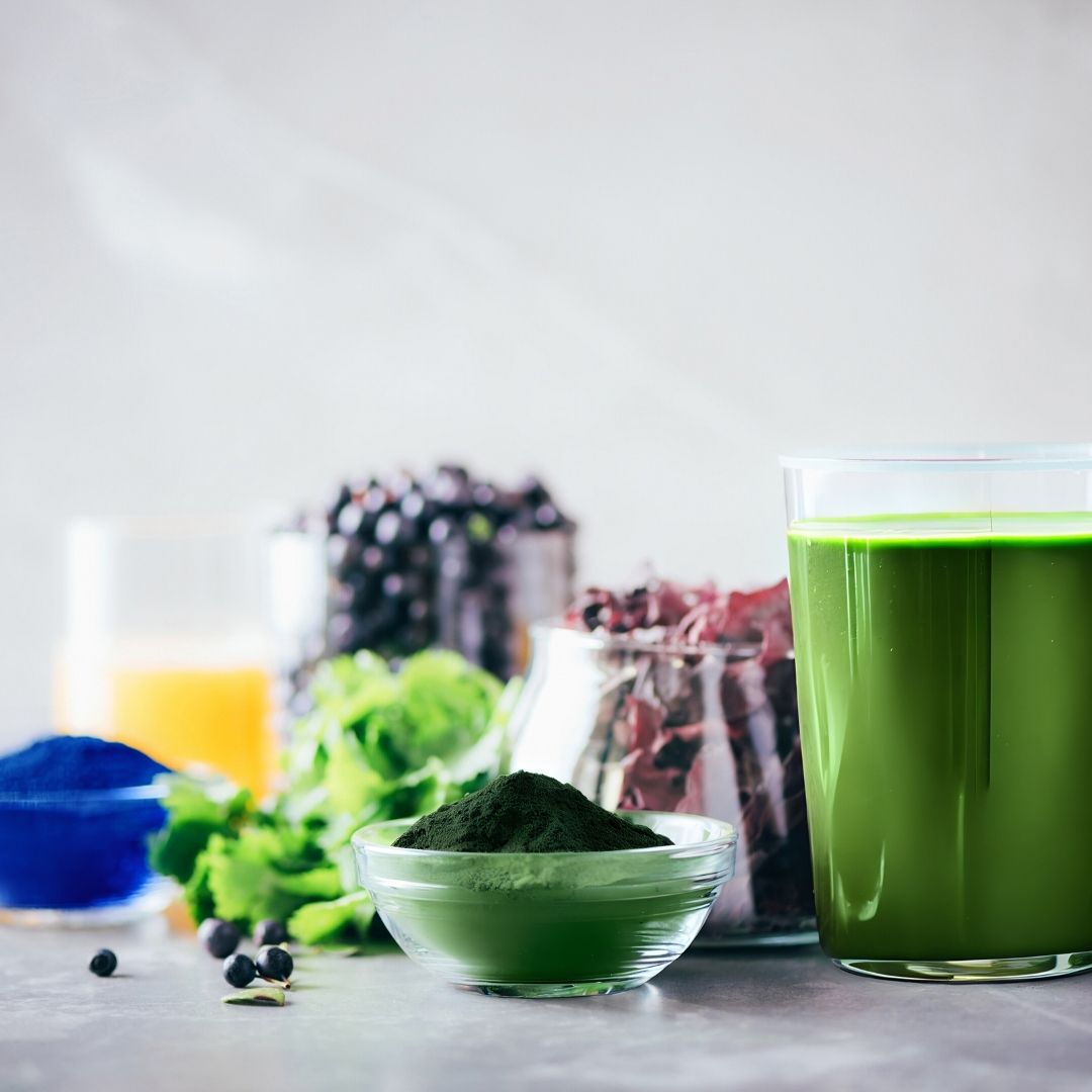 Green smoothie in a glass with ingredients like spinach, berries, and a bowl of green powder on a light background.