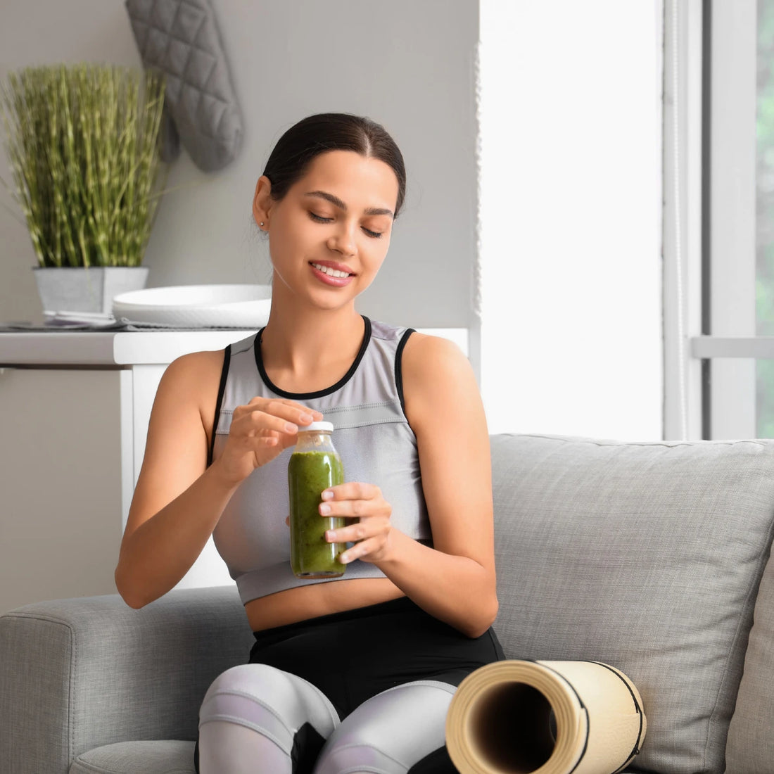 Woman sitting on a couch holding a green smoothie jar in a home setting.