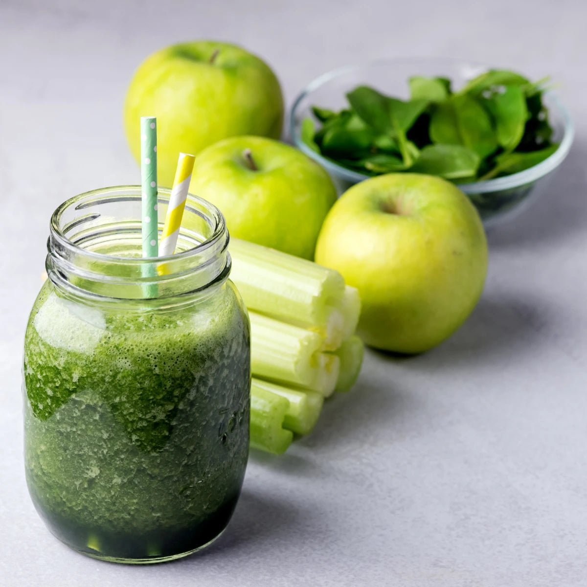 Green smoothie in a jar with a straw, surrounded by green apples, celery, and spinach on a light gray background.