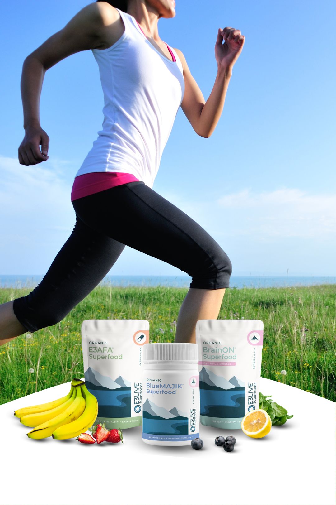 A woman in athletic wear is running outdoors on a grassy field near the ocean under a clear blue sky. In the foreground are three E3Live® Superfoods products: E3AFA® capsules, BlueMAJIK® powder, and BrainON® Singles powder. Surrounding the products are fresh fruits and greens including bananas, strawberries, blueberries, spinach, and lemon, symbolizing natural energy and wellness.