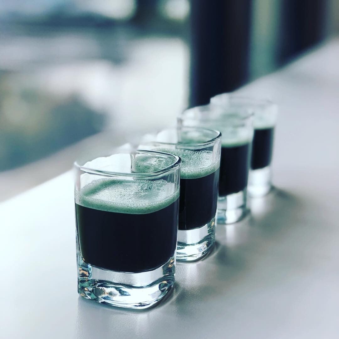 Four shot glasses with E3Live liquid and a green foam layer on a reflective surface.