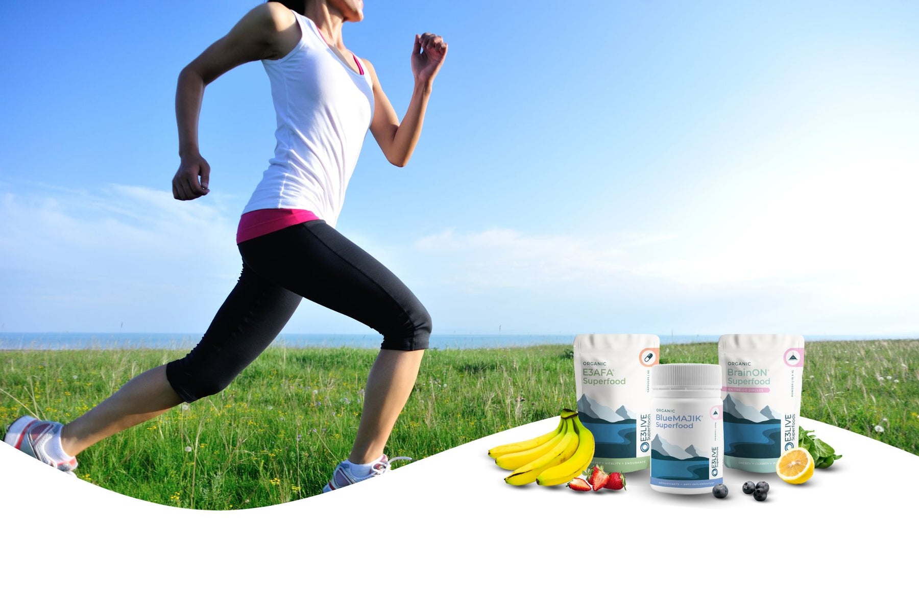 A woman in athletic wear is running outdoors on a grassy field near the ocean under a clear blue sky. In the foreground are three E3Live® Superfoods products: E3AFA® capsules, BlueMAJIK® powder, and BrainON® Singles powder. Surrounding the products are fresh fruits and greens including bananas, strawberries, blueberries, spinach, and lemon, symbolizing natural energy and wellness.