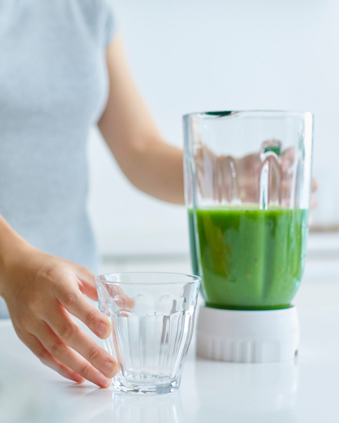 Person holding a glass next to a blender filled with green juice on a white surface.
