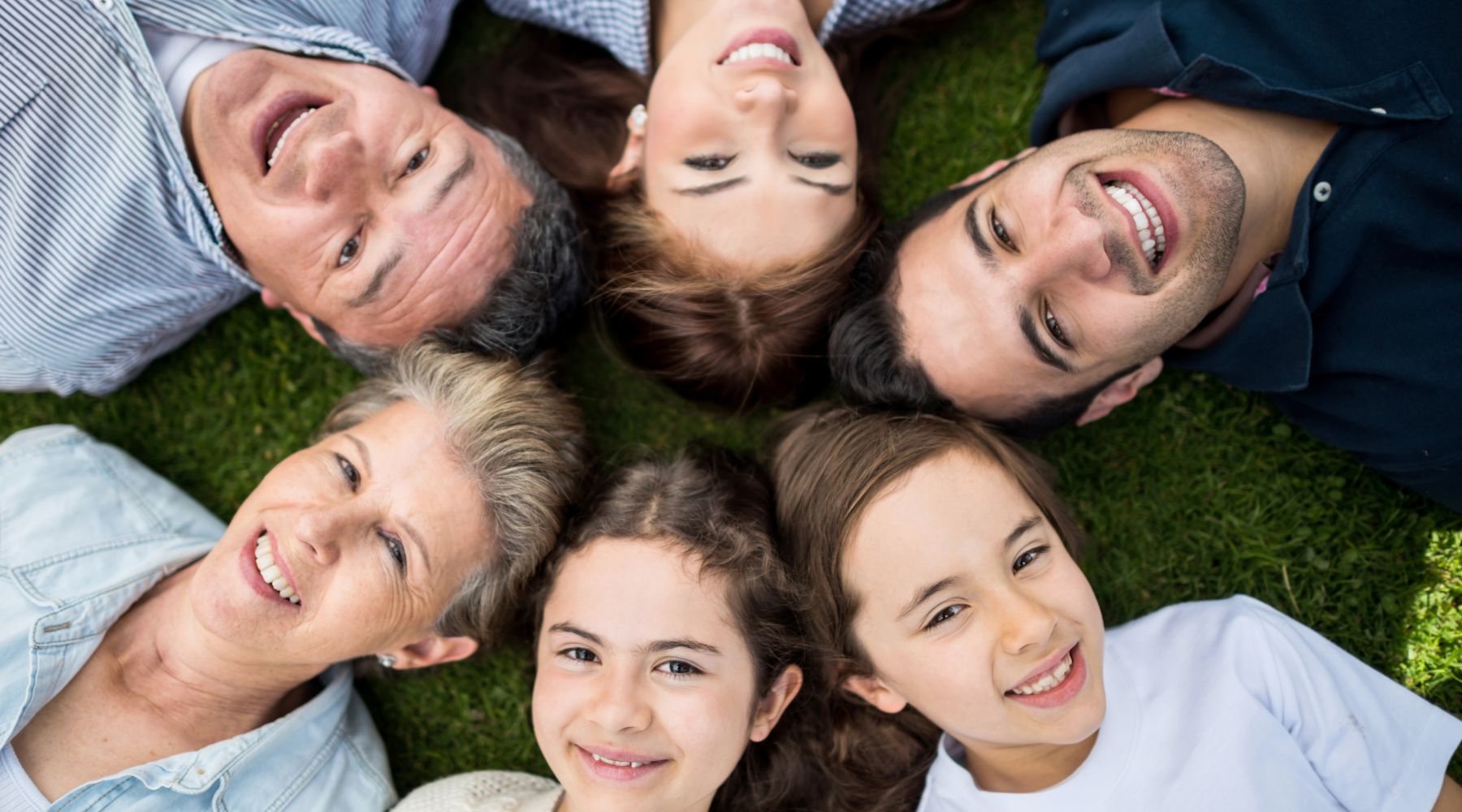 Group of smiling family members of various ages lying on grass in a circular arrangement, heads together, showcasing joy and togetherness. The image highlights a multigenerational family with adults, seniors, and children in a vibrant outdoor setting.