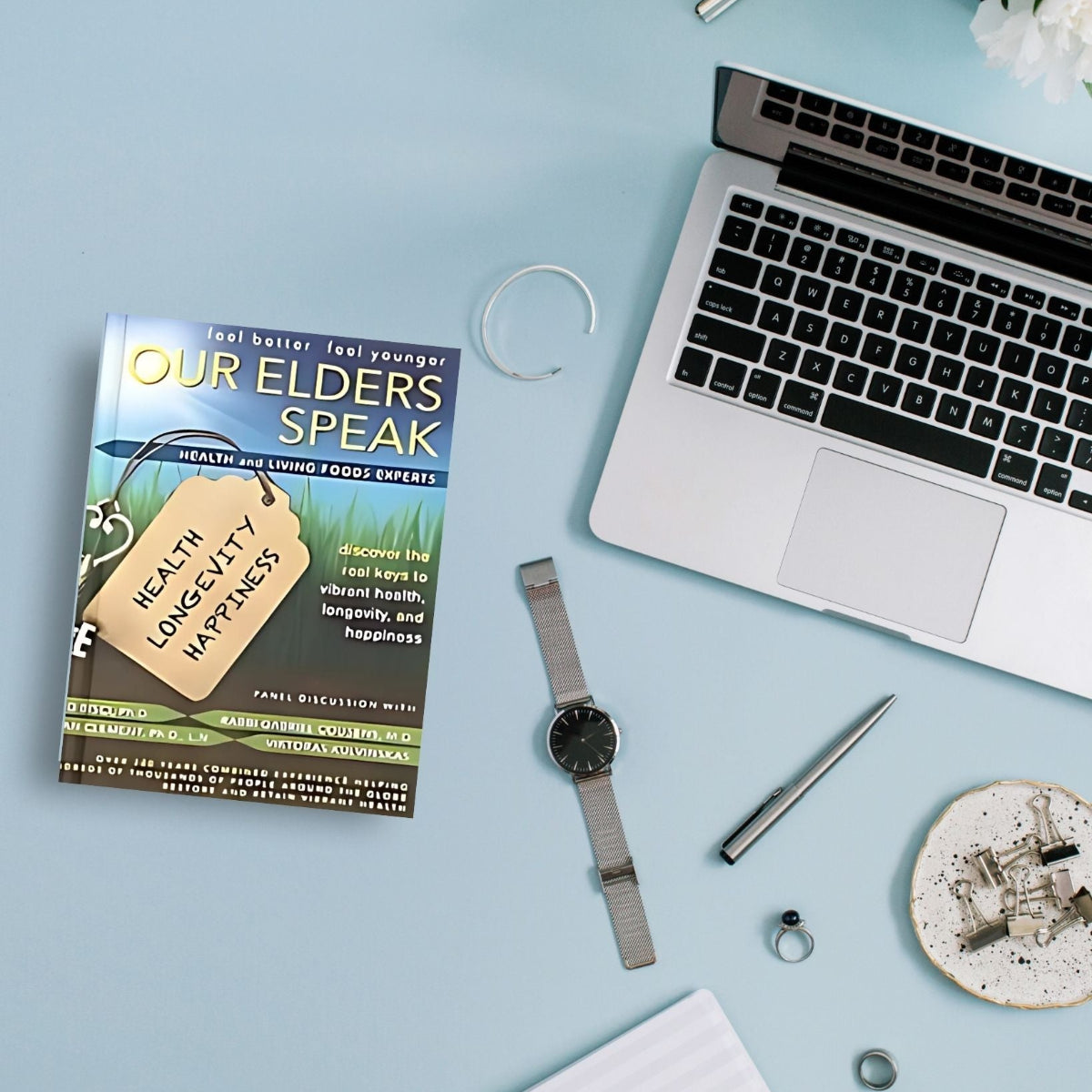 Book titled 'Our Elders Speak' on a light blue surface with a laptop, watch, pen, and other items.