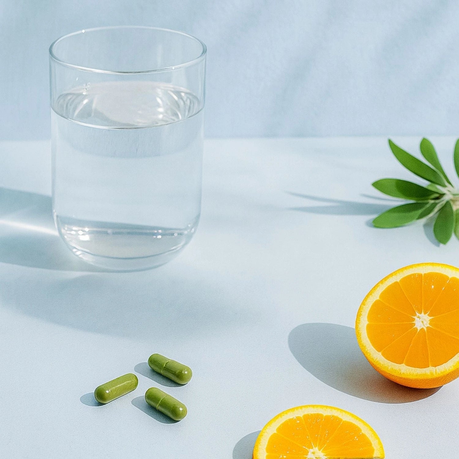 Glass of water with green RenewME capsules, half an orange, and leaves on a light blue background