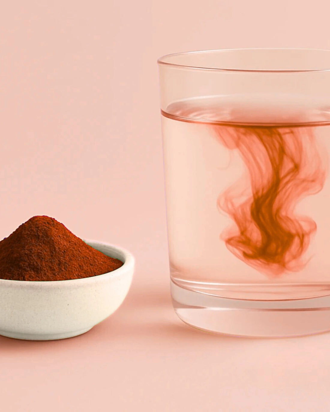 Astaxanthin powder in a small bowl next to a glass of water with red powder swirls on a pink background