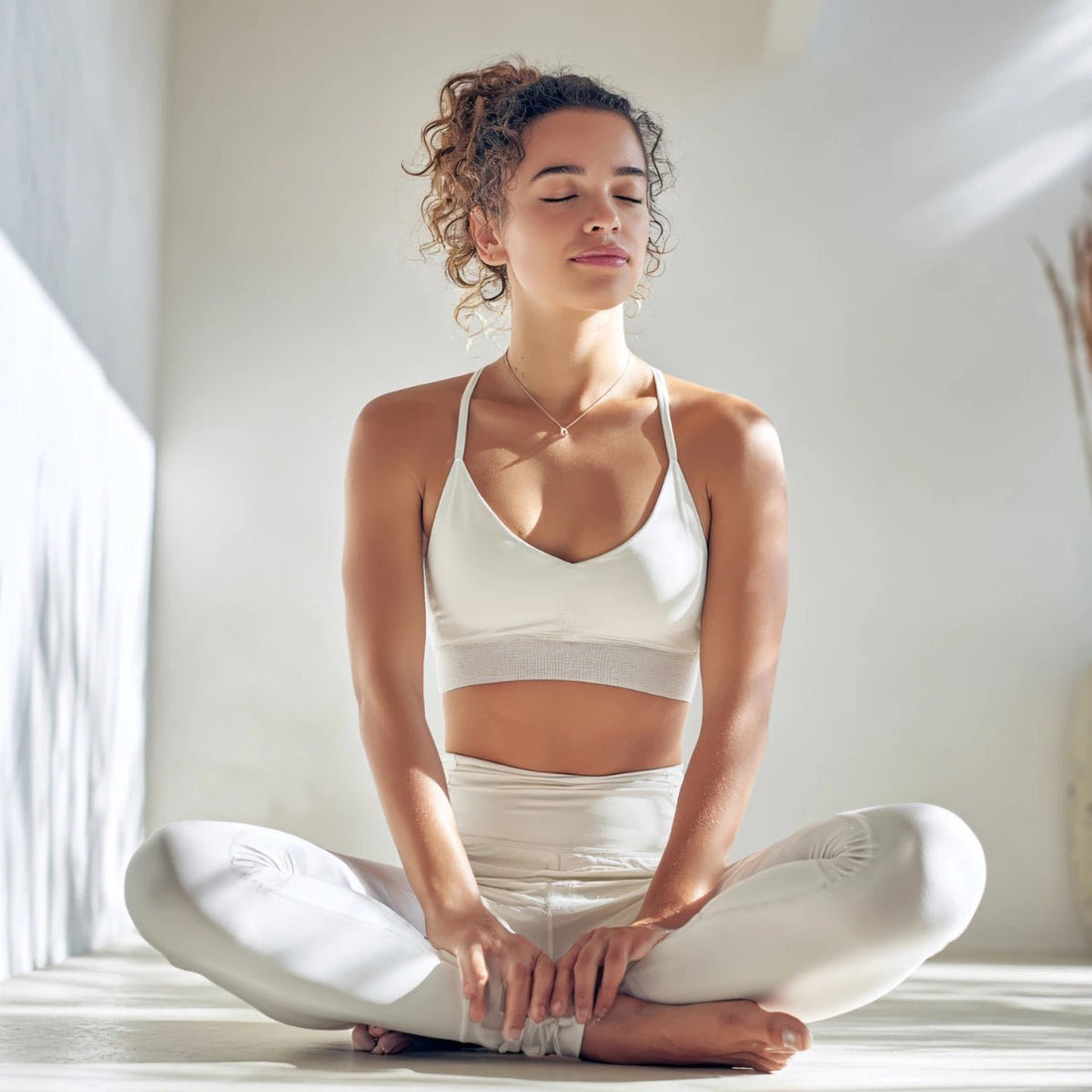 Woman in a white sports bra and leggings sitting in a yoga pose with eyes closed.