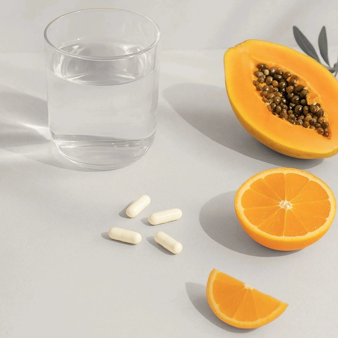 Glass of water, half-cut papaya, half-cut orange, and capsules on a light gray background