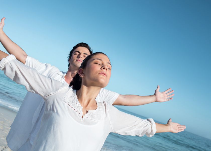 Man and woman in white clothing standing on a beach with arms outstretched, eyes closed, and faces lifted toward the sky — expressing a sense of calm, balance, and wellbeing.