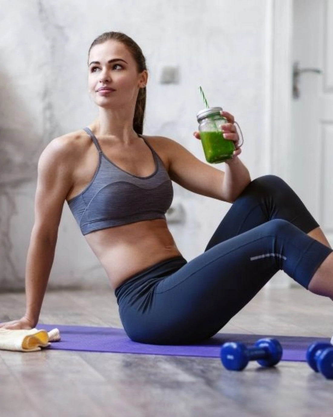 Woman in workout attire holding a green smoothie with exercise equipment nearby.