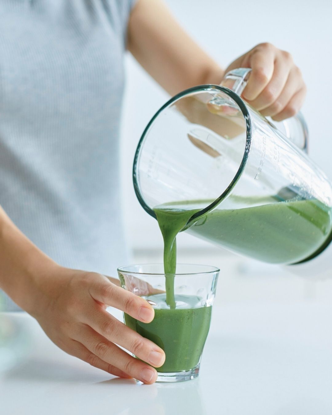 Person pouring green juice from a pitcher into a glass on a light background