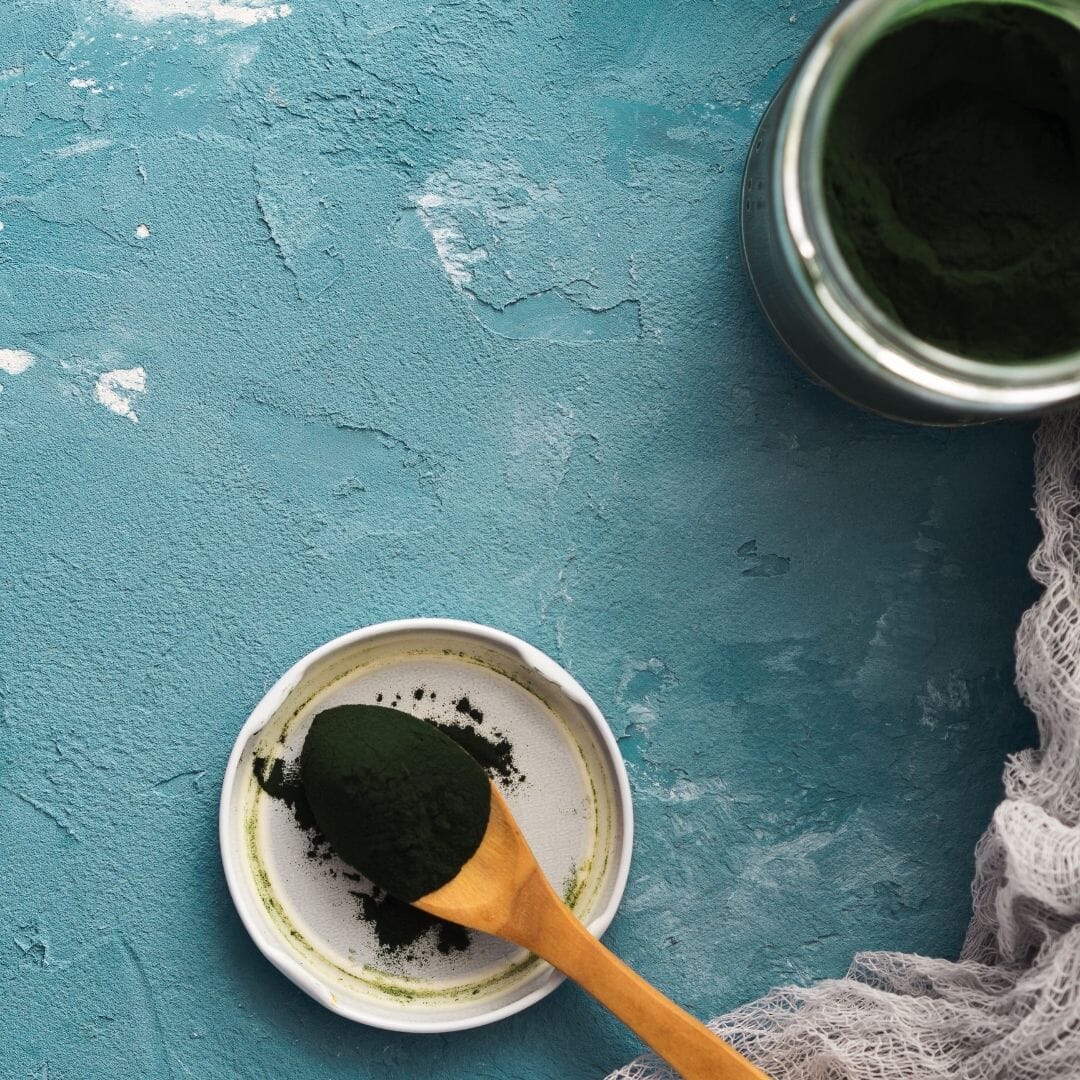 Green powder in a bowl with a wooden spoon on a textured blue surface